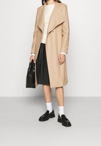 Beige uldfrakke med draperet revers og enkelt knappelukning, parret med en hvid top, plissee-sort nederdel, sort taske og sorte sko.