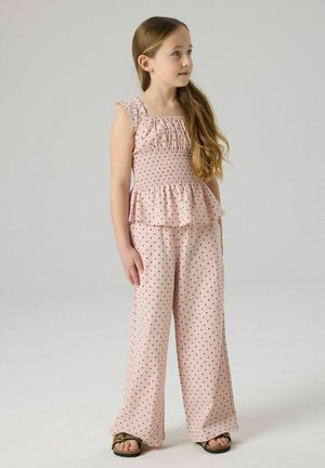 Mladá dívka s dlouhými vlasy v růžovém tílku s puntíky a odpovídajících širokých kalhotách, stojí a dívá se stranou.