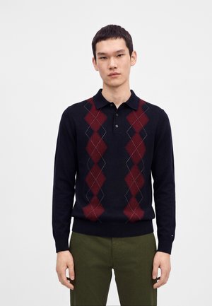 Jonge man die een marineblauwe polo met lange mouwen draagt met een rood argyle-patroon en olijfgroene broek, staand tegen een egale lichte achtergrond.