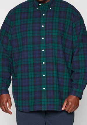 Muž v tmavě zelené a námořnicky modré kostkované košili s dlouhými rukávy a modrých kalhotách, stojící před jednobarevným pozadím.