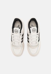 White sneakers with black stripes, made of leather and textile. Features perforated toe box, flat laces, and logo on the tongue. Classic design.