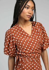 Robe portefeuille à pois marron avec des manches courtes bouffantes, un décolleté en V et une taille nouée. Fabriquée dans un tissu lisse avec un léger éclat.