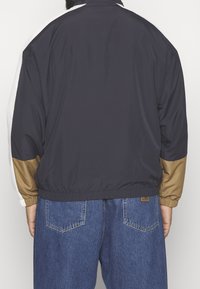 Marinblå vindjacka med elastiska ärmslut, en beige detalj på ärmarna och en avslappnad passform, tillsammans med blå denimjeans.