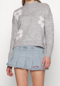 Maglione grigio chiaro con motivi floreali rosa, scollo rotondo e vestibilità rilassata. Abbinato a una gonna a pieghe in denim con ricamo "BDG JEANS".