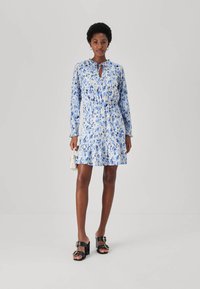 Langärmliges, blaues Blumenkleid mit gebundenem Ausschnitt, tailliertem Bund und rüschengesäumtem Saum; kombiniert mit schwarzen Blockabsatzsandalen und einer kleinen Tasche.
