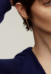 Pendiente dorado con una gema ámbar en forma de cuadrado y pequeños acentos claros, conectado a un diseño de gota en cadena. Lleva un peinado corto.