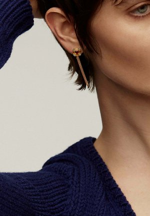 Pendiente dorado con una gema ámbar en forma de cuadrado y pequeños acentos claros, conectado a un diseño de gota en cadena. Lleva un peinado corto.
