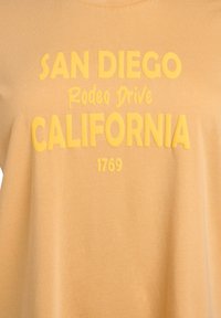 Camiseta gráfica amarilla con la inscripción "SAN DIEGO Rodeo Drive CALIFORNIA 1769" en una fuente audaz y gruesa sobre una tela suave y texturizada.