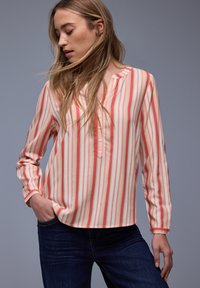 Femme blonde aux cheveux lâchés portant un chemisier à manches longues à rayures verticales corail, pêche et blanc, et un jean bleu foncé, main dans la poche.