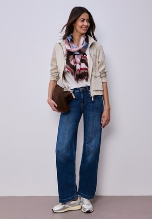 Femme souriante, portant une veste légère, une écharpe à motifs, une chemise blanche, un jean bleu à jambes larges, des baskets blanches, et tenant un sac marron.