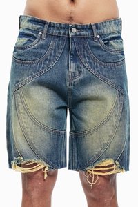 Denim shorts dengan ujung yang dipotong, menampilkan warna biru pudar, detail jahitan melengkung, dan pinggiran yang terurai, menonjolkan kain yang berpola.