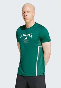 Camiseta deportiva verde de manga corta, con la marca "ADIDAS" y un panel lateral blanco en contraste. Textura suave y diseño ajustado.