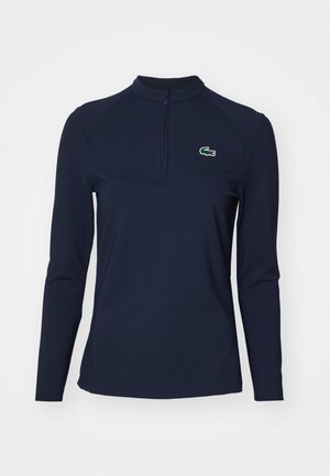 Polo à manches longues bleu marine avec col zippé. Présente une texture côtelée et un petit logo vert et rouge sur le côté gauche de la poitrine.