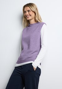 Lavendelstickad ärmlös tröja över en vit långärmad skjorta, i kombination med marinblå byxor. Slät textur, hög krage design.