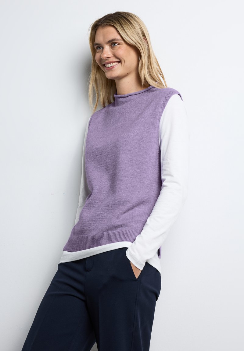 Lavendelstickad ärmlös tröja över en vit långärmad skjorta, i kombination med marinblå byxor. Slät textur, hög krage design.