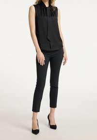 Femme portant un haut sans manches noir avec un nœud au cou, un pantalon noir slim et des chaussures à talons hauts noires, se tenant devant un fond uni.