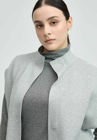 Gray, lightweight jacket with a rounded collar and button closure, layered over a dark gray, sparkly turtleneck. Smooth texture.