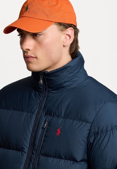 Giacca puffer blu navy con colletto alto e logo rosso. Accompagnata da un berretto arancione, caratterizzato da una visiera strutturata e dettagli ricamati.