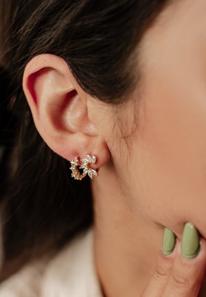 Close-up of ear wearing a delicate gold hoop earring with clear marquise and round gemstones, hand with green nail polish touching jawline.