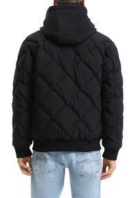 Giacca a vento nera trapuntata con cappuccio, polsini e orlo a costine. Presenta cuciture diagonali e una vestibilità rilassata. Indossata con jeans blu chiaro.