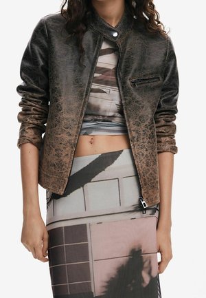 Vrouw draagt een verweerde bruine leren jas met opgerolde mouwen over een patroonrijke crop top en een hoge taille rok die de buik laat zien.