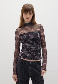 Top largo negro y transparente con flores, de manga larga y cuello alto, con detalle fruncido en la cintura, combinado con pantalones oscuros de corte holgado.