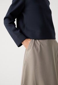 Persona vestida con una blusa de manga larga azul marino y una falda beige, con una mano casualmente colocada en el bolsillo de la falda, contra un fondo claro y liso.