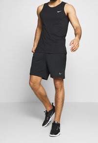 Mies, który ma na sobie czarną bezrękawnik Nike, czarne szorty i czarne sneakersy, stoi na tle jasnym.