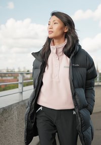 Juodas pūkinis švarkas su aukštu apykakle, dėvimas virš šviesiai rožinio megztinio. Juodos kelnės su baltomis užtrauktukų kišenėmis, lauko aplinka su debesimis.