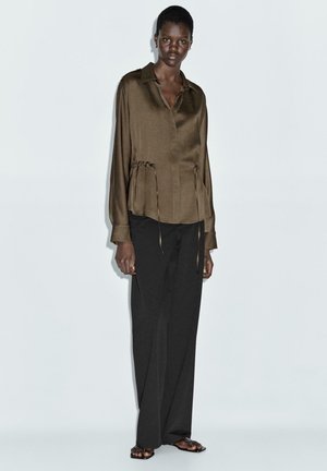 Braune Satin-Hemdbluse mit langen Ärmeln, Kragen und Kordelzugdetail an der Taille, kombiniert mit lockeren schwarzen Hosen und flachen Sandalen.