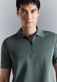 Joven con cabello corto y negro que lleva una camisa polo verde de manga corta con cremallera frontal, de pie frente a un fondo gris liso.