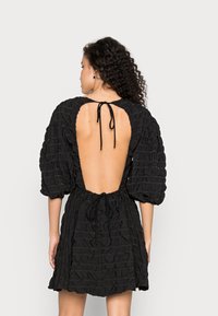 Vestido preto feito de tecido texturizado com mangas bufantes, costas abertas seguradas por um laço, e um ajuste relaxado, apresentando uma cintura franzida.