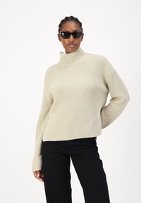Beige ribbet turtleneck genser med lange ermer, som har en avslappet passform. Sammen med svarte bukser og kantete svarte solbriller.