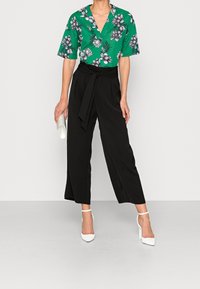 Blusa floral verde con escote en forma de V, combinada con pantalones anchos negros. La modelo sostiene un bolso de mano blanco y lleva zapatos de tacón blanco.