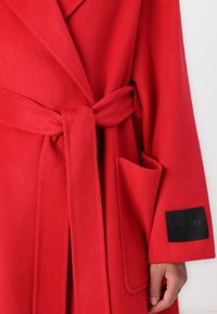 Roter Fleece-Bademantel mit schalähnlichem Kragen, abnehmbarem Gürtel, seitlichen Taschen und einem schwarzen Markenpatch am Ärmel. Weiche Textur.