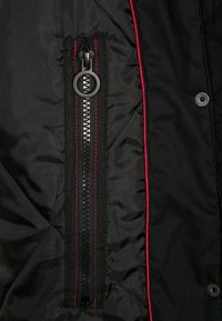 Tissu matelassé noir avec fermeture éclair noire verticale bordée de coutures rouges et tirette de fermeture éclair en métal circulaire, à côté d'une couture bordée de rouge et d'un bouton-pression.