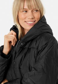 Giacca puffer nera con colletto alto, sezioni spesse trapuntate e chiusura frontale con zip. Il tessuto appare liscio e resistente all'acqua.