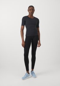 Schwarzes, eng anliegendes Oberteil mit kurzen Ärmeln, kombiniert mit schwarzen Leggings und hellblauen Sportschuhen. Glattes Material, schlichtes Design und maßgeschneiderte Passform.