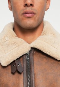 Primo piano del volto inferiore di una persona che indossa una giacca di pelle marrone con un colletto in shearling beige e una zip metallica frontale.