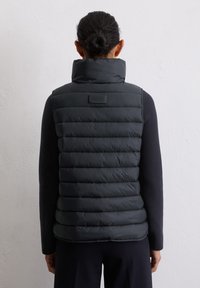 Femme aux cheveux foncés en chignon, portant un gilet sans manches matelassé noir par-dessus un haut à manches longues, se tenant devant un mur blanc uni.