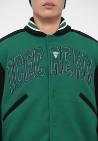 Giacca varsity verde con maniche nere, colletto a coste con strisce bianche ed ricamo stilizzato "ICECREAM" sul petto.