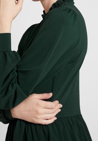 Tmavě zelené, strukturované šaty s dlouhými nabíranými rukávy a volánkovým výstřihem; nařasený pas pokračuje do plisované sukně.