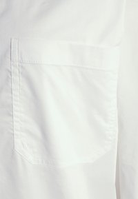 Tecido de camisa branca com uma textura suave. Apresenta um único bolso no peito com uma borda superior curva e costura visível.