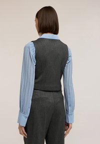 Grauer maßgeschneiderter Weste aus strukturiertem Stoff, kombiniert mit einer hellblauen Bluse mit vertikalen Nadelstreifen und geknöpften Manschetten.
