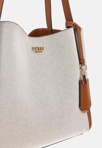Sac à main argenté avec une surface texturée, des accents en cuir marron et un logo doré. Il dispose de doubles poignées et d'une petite étiquette attachée.