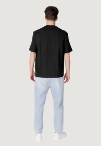 Maglietta nera a maniche corte con effetto texture e colletto tondo, vista da dietro. Abbinata a pantaloni larghi azzurri e sneakers bianche.