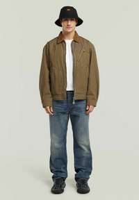 Veste bomber marron avec un col contrastant, t-shirt blanc, jeans bleus et chapeau seau noir. Elle possède des poches et un détail de fermeture éclair.