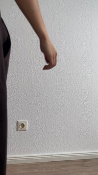 Vue partielle d'une main atteignant un mur texturé blanc. Le sol est en bois, avec une prise électrique visible près de la plinthe.