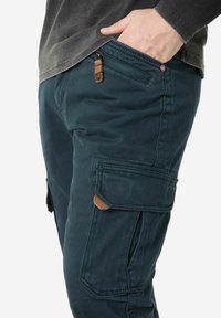 Dark green cargo pants made of textured fabric featuring multiple pockets and a contrasting brown leather accent on the waistband.