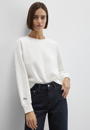 Woman with straight brown hair wears a white long-sleeve top with "M" and heart on cuff, paired with dark blue jeans, standing against a white wall.
